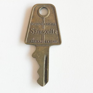 May include: Vintage brass key with the text "Samsonite" and the cities "Denver" and "Detroit" printed on it. The key has a rectangular head with a circular hole and a jagged edge on the blade.
