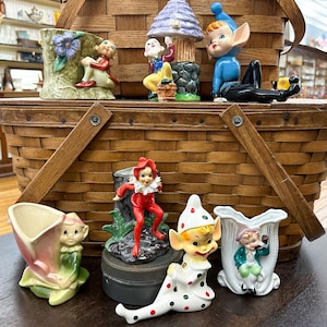 May include: A collection of vintage elf figurines, some holding flowers, others standing on tree stumps. The figurines are made of ceramic and painted in bright colors, including red, green, blue, and white. The figurines are arranged in a wicker basket.