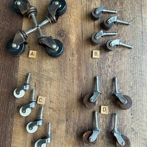 May include: An assortment of antique furniture casters, including four different styles. The casters are made of metal and wood, with black or white wheels. The casters are arranged on a wooden surface.