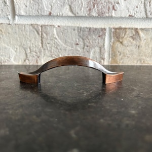 May include: A copper-toned cabinet pull with a curved arch design. The pull is made of metal and has a smooth, polished finish.