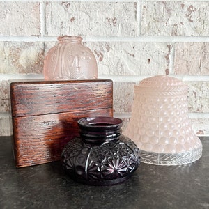 May include: An assortment of antique glass and wooden items. Includes a dark purple inkwell with a starburst pattern, two pink glass shades, and a small wooden box with a hinged lid. The items are arranged on a dark surface.
