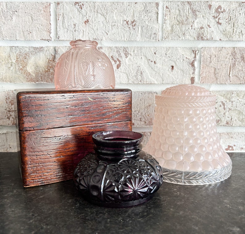 May include: An assortment of antique glass and wooden items. Includes a dark purple inkwell with a starburst pattern, two pink glass shades, and a small wooden box with a hinged lid. The items are arranged on a dark surface.