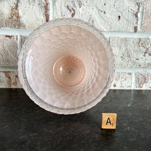 May include: A clear glass vase with a textured, honeycomb-like pattern on the inside. The vase has a light pink hue and a decorative rim. The vase is sitting on a dark surface with a brick wall in the background.