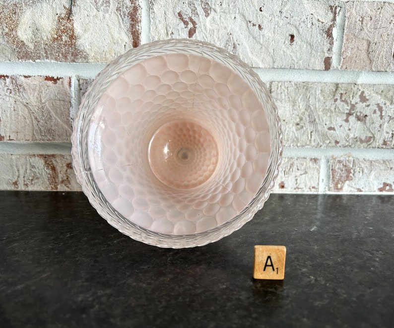 May include: A clear glass vase with a textured, honeycomb-like pattern on the inside. The vase has a light pink hue and a decorative rim. The vase is sitting on a dark surface with a brick wall in the background.