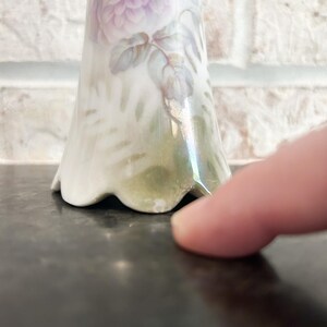Puede incluir: Un jarr&oacute;n de porcelana blanca con base acampanada y dise&ntilde;o floral pintado. El dise&ntilde;o presenta una gran rosa de color p&uacute;rpura claro con hojas y tallos verdes. El jarr&oacute;n tiene un borde festoneado.