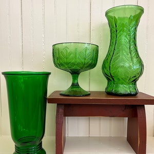 May include: Three green glass vases of different sizes and shapes, with a leafy pattern, sitting on a small wooden stool.