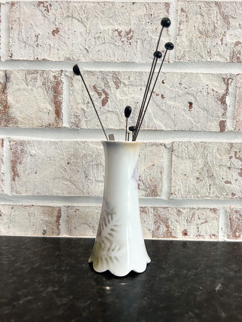 Puede incluir: Un peque&ntilde;o jarr&oacute;n de porcelana blanca con base festoneada, adornado con un dise&ntilde;o floral en tonos morados y verdes. El jarr&oacute;n contiene varios tallos decorativos finos con punta negra. El fondo es una pared de ladrillo blanco.