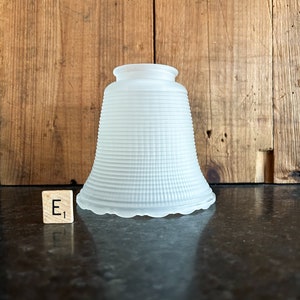 May include: A frosted glass lampshade with a ribbed design. The lampshade has a flared shape with a scalloped edge at the bottom. The background is a wooden surface.