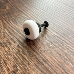 May include: White ceramic knob with a black center and a black metal screw. The knob is on a wood surface.