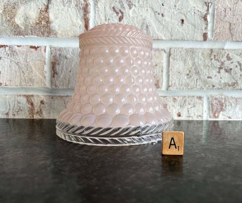 May include: A light pink glass lampshade with a textured, bubble-like pattern. The base has a decorative, clear glass trim. A wooden tile with the letter "A" and the number "1" is next to the lampshade.