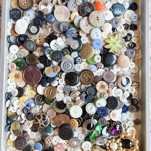 May include: A large collection of assorted buttons in various shapes, sizes, and colors, including round, star-shaped, and decorative styles, scattered across a rectangular metal tray. The buttons feature diverse materials, textures, and finishes.