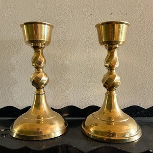 May include: Two brass candlesticks with a twisted stem design. The candlesticks have a wide base and a cup-shaped top to hold candles. The brass has a polished finish, reflecting light. The candlesticks are displayed on a black surface.