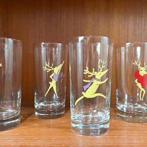 May include: Four clear drinking glasses with cartoon reindeer designs. Each glass features a different reindeer character in various poses and outfits, with the names of the reindeer printed on the glasses.