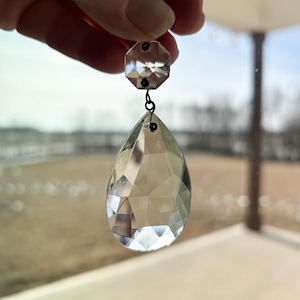 May include: A clear glass teardrop-shaped crystal prism with a smaller octagonal crystal prism at the top. The prisms are connected by a thin metal wire.