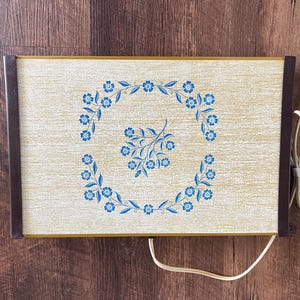 May include: Rectangular electric warming tray with a cream-colored surface and a blue floral design. The tray has dark brown edges and a gold trim. A white power cord extends from the side. The tray is approximately 18 inches long and 12 inches wide.