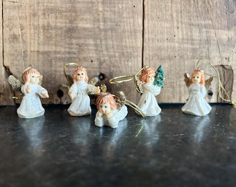 May include: A collection of small angel ornaments with gold wings, peach-colored hair, and white gowns. One angel holds a small green Christmas tree. The ornaments are displayed against a rustic wooden background, perfect for holiday decor.