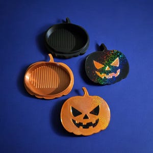 May include: Four pumpkin-shaped Halloween decorations. One is black, one is orange, and two are orange with a black face. The black pumpkin has a rainbow glitter effect. The orange pumpkins have a carved face.