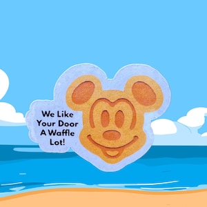 May include: A Mickey Mouse-shaped waffle design with the text "We Like Your Door A Waffle Lot!" against a blue sky and beach background. The waffle is light brown with a smiling face.
