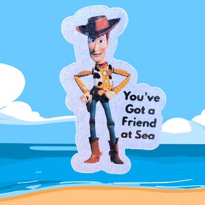 May include: A sticker of the Toy Story character Woody, in a cowboy outfit, with the text "You've Got a Friend at Sea". The background shows a beach and sea, with a blue sky.