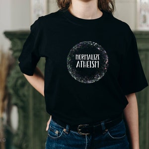 May include: Black t-shirt with the text "Normalize Atheism" inside a glittery circle. The shirt is a crew neck style and is paired with blue jeans. The overall aesthetic is casual and modern.