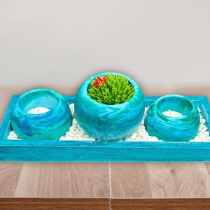 May include: A decorative teal wooden tray holds three spherical teal candle holders with white candles. The center holder contains a small green plant with a red flower. White pebbles fill the tray.