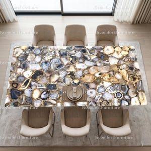 May include: A rectangular dining table with a unique agate stone top in shades of blue, gold, and white. The table is surrounded by six beige upholstered chairs. A place setting with plates, silverware, and a napkin is visible on the table.