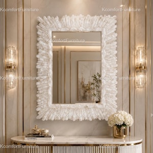 May include: A rectangular white mirror with a textured, crystal-like frame hangs above a marble-topped console. The console features a gold trim and holds a gold vase with white flowers, books, and decorative items. Two wall sconces flank the mirror.