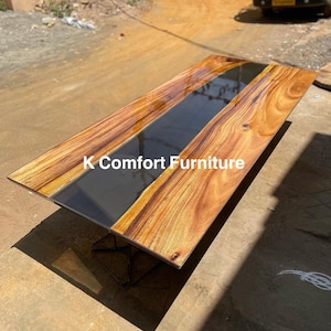 Op de afbeelding: Een lange, rechthoekige houten tafel met een zwarte epoxyhars inlay. De tafel heeft een natuurlijke houtfinish en wordt ondersteund door een zwart metalen frame. De tafel is gelabeld "K Comfort Furniture".