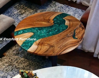 May include: Round wooden coffee table with a green epoxy resin river design. The table has a natural wood grain finish and a black metal base. "K Comfort Furniture" is visible on the table.