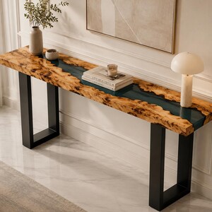 May include: A long console table featuring a live-edge wood slab with a central dark blue resin river inlay, supported by two black rectangular metal legs. The surface holds a ceramic vase with greenery, a candle, two stacked books, and a table lamp.