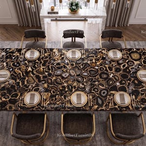 Puede incluir: Una mesa de comedor con una superficie de piedra de ágata negra y dorada, preparada para ocho comensales. La mesa está rodeada de sillas grises oscuras con marcos dorados, sobre una alfombra gris. La habitación tiene grandes ventanas y cortinas neutras.