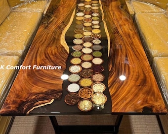 May include: A wooden table with a black epoxy resin river running down the center. The river is filled with small bowls of various grains and seeds. The table is made of dark wood with a natural finish. "K Comfort Furniture" is printed on the table.