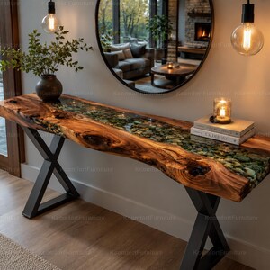 Puede incluir: Una mesa consola de madera con borde natural y un diseño de río central relleno de piedras verdes y resina transparente. La mesa tiene un acabado de veta de madera natural y se apoya en dos patas de metal negro en forma de X. Dos libros y una vela.