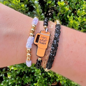 May include: A set of three bracelets featuring gold beads, black beads, and white glitter beads. The bracelets are stacked on a person's wrist. A charm with the text "I can't adult today" is attached to the bracelets.