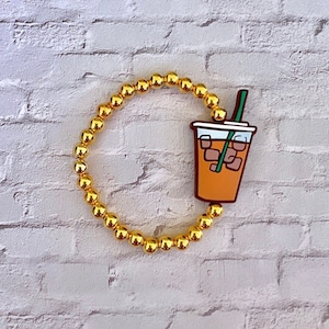 May include: A gold beaded bracelet with a charm of an iced coffee cup with a straw. The charm is orange, brown, and green.