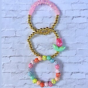 May include: Three bracelets with gold beads and colorful accents. The top bracelet has pink beads with glitter, the middle bracelet has gold beads, and the bottom bracelet has pastel colored beads with silver accents. A small pink and green flower charm is attached to the middle bracelet.