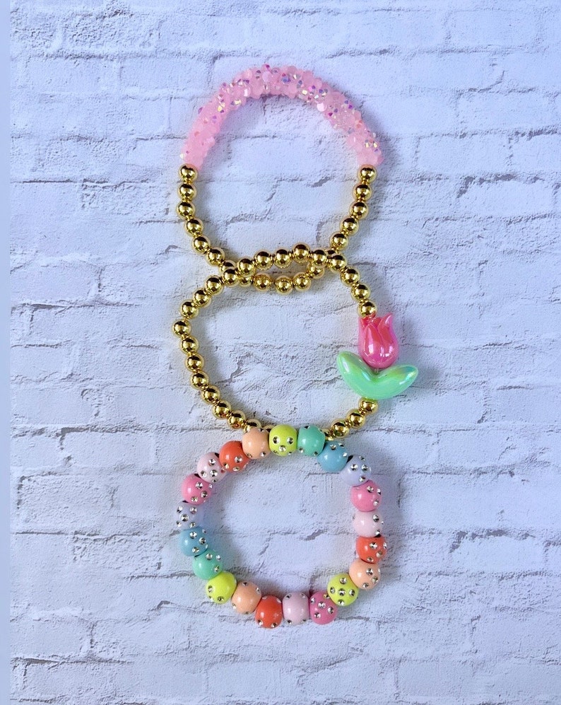 May include: Three bracelets with gold beads and colorful accents. The top bracelet has pink beads with glitter, the middle bracelet has gold beads, and the bottom bracelet has pastel colored beads with silver accents. A small pink and green flower charm is attached to the middle bracelet.