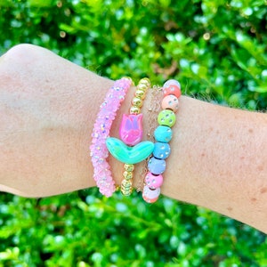 May include: A set of four colorful beaded bracelets. The bracelets feature a pink and green flower charm, gold beads, and pastel-colored beads with rhinestones.