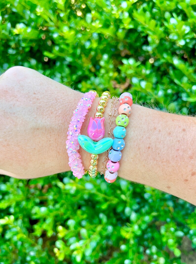 May include: A set of four colorful beaded bracelets. The bracelets feature a pink and green flower charm, gold beads, and pastel-colored beads with rhinestones.