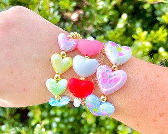 Peut inclure: Un bracelet avec une chaîne dorée et des perles en forme de cœur colorées. Les cœurs sont décorés de différents motifs et designs, notamment des pois, des fleurs et le mot "love".