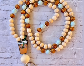 Puede incluir: Un cordón de cuentas de madera con un esquema de colores azul claro y marrón. El cordón tiene un dije de una taza de café helado de dibujos animados con una pajita.