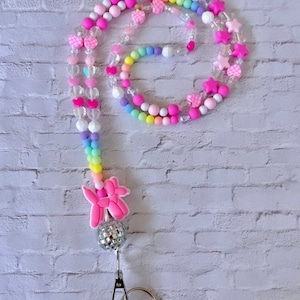 May include: A colorful beaded lanyard with pink hearts, stars, and checkered patterns. The lanyard has a pink balloon animal charm and a silver disco ball charm. The lanyard is attached to a silver key ring.