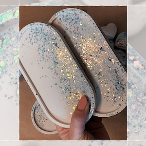 Peut inclure: Deux plateaux ovales en résine blanche avec une finition scintillante irisée. Les plateaux sont empilés l'un sur l'autre.