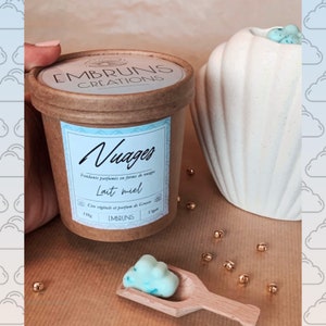 Op de afbeelding: Een bruine container met een wit label waarop "Nuages" en "Lait miel" staat, met een blauwe en witte wolkvormige was smelt op een houten lepel. De container is gelabeld met "Embruns Creations".