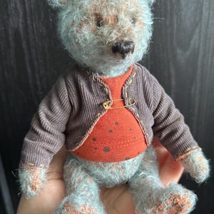 May include: A handmade teddy bear with a light blue and brown color scheme. The bear is wearing a brown corduroy jacket secured with a safety pin over an orange shirt. The bear's paws and feet have a textured, patterned appearance.