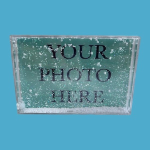 May include: A clear, rectangular photo frame with the words "YOUR PHOTO HERE" in black. The frame has a teal background with white speckles, resembling snow. The frame is set against a bright blue backdrop.