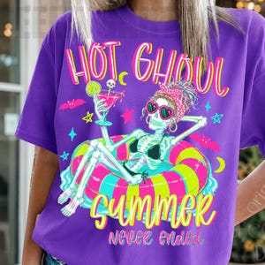 May include: Purple t-shirt with a skeleton graphic wearing sunglasses and a bikini, lounging on a colorful inflatable ring. The shirt features the text "HOT GHOUL SUMMER never ended" in bright colors, with stars and bats.