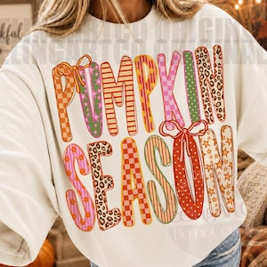 May include: An off-white crewneck sweatshirt featuring the text PUMPKIN SEASON in large, multi-colored letters. Each letter displays a unique pattern, including leopard print, checkered, striped, floral, and star designs, accented with orange bows.