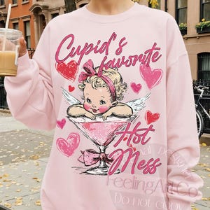 May include: Pink sweatshirt with a graphic of a baby cupid in a martini glass, surrounded by hearts. The text "Cupid's favorite Hot Mess" is displayed in pink script. The sweatshirt is a light pink color.