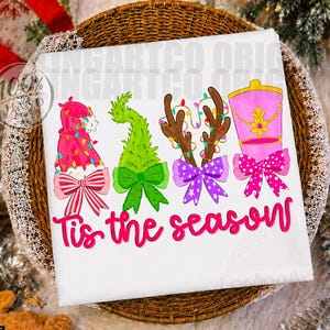 May include: A white t-shirt featuring a graphic print of four holiday-themed icons: a pink hat with string lights, a green fuzzy shape, brown reindeer antlers with lights, and a pink drum, all with bows. The text 'Tis the season' is in pink.